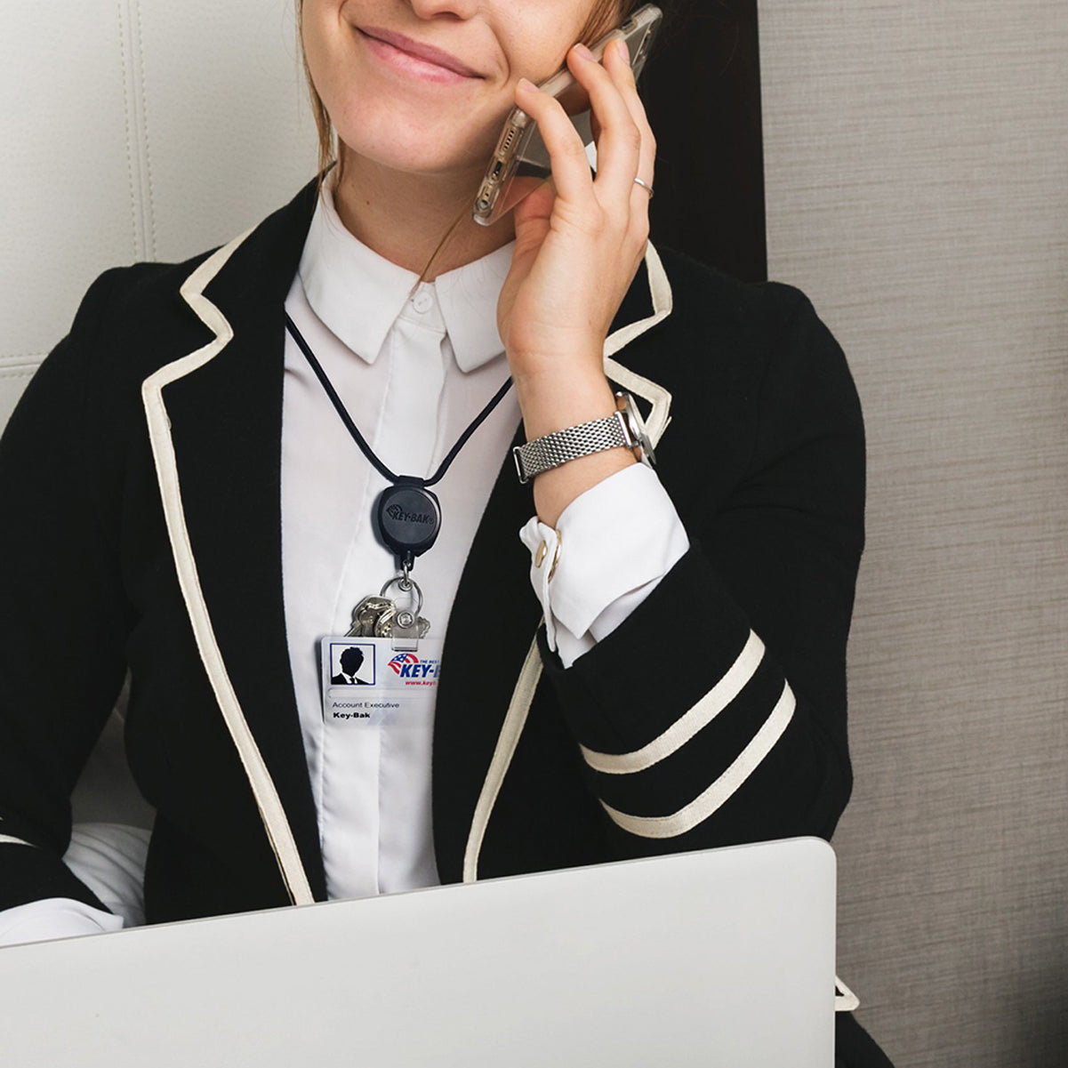 A person in a black blazer with white trim and wearing an ID badge attached to a Key-Bak Heavy Duty Paracord Lanyard With Sidekick Retractable ID Badge & Key Reel (0KB1-0A44) talks on the phone while using a laptop.