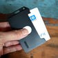 A hand holds a 2 Sided Rigid Vertical MultiCard Badge Holder - Hard Plastic Multiple ID Card Holder (1840-308X), containing a white ID card labeled "HID." The ID card partially slides out of the holder.