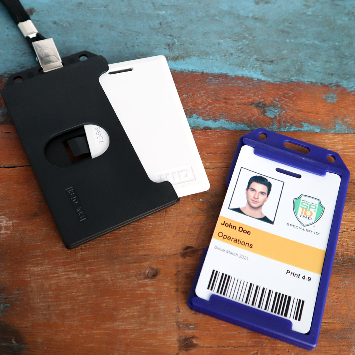 Close-up of two ID badges on a wooden surface. One ID is white in a black rigid plastic holder, and the other badge is inside a purple holder displaying a photo, text "John Doe Operations", and a company logo. The 2 Sided Rigid Vertical MultiCard Badge Holder - Hard Plastic Multiple ID Card Holder (1840-308X) ensures secure placement for multiple essential IDs.