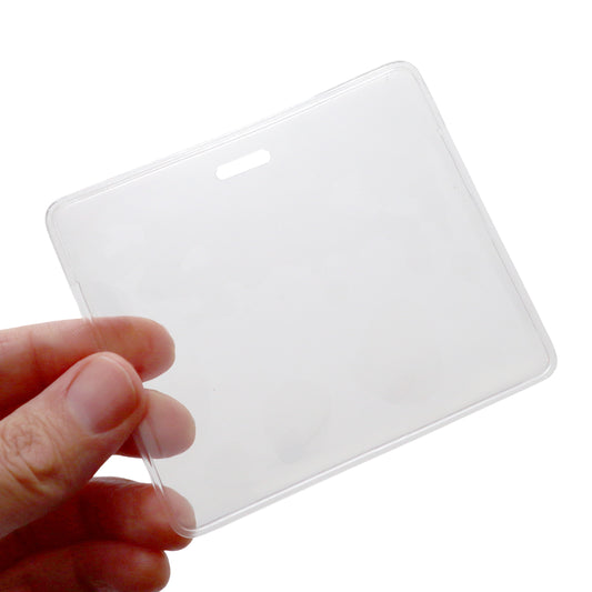 A hand is holding a Horizontal Badge Buddy Holder (P/N 1840-5014) with a transparent, empty plastic enclosure and a slot at the top for attaching a lanyard, featuring a sturdy vinyl pocket for reliable protection.