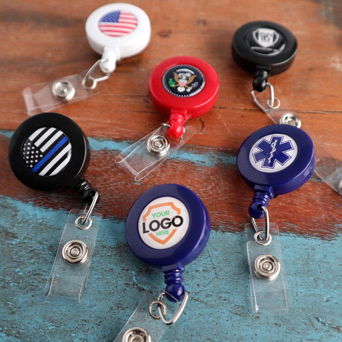 Various Custom Printed Retractable Badge Reels With Belt Clip - Personalize with Your Brand Logo on a wooden surface, featuring designs such as an American flag, medical symbol, and customizable logo with full color graphics to promote brand awareness.