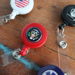 A close-up of Custom Printed Retractable Badge Reels With Belt Clip - Personalize with Your Brand Logo with various designs, including an eagle emblem on a red holder, placed on a wooden surface. Perfect for promoting brand awareness, these retractable holders feature full-color graphics.