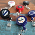 Assorted Custom Printed Retractable Badge Reels With Belt Clip - Personalize with Your Brand Logo lie on a wooden surface. Some show emblems, while one has a placeholder for a custom logo to promote brand awareness. Featuring retractable cords and metal clip attachments, they also showcase vibrant full color graphics.