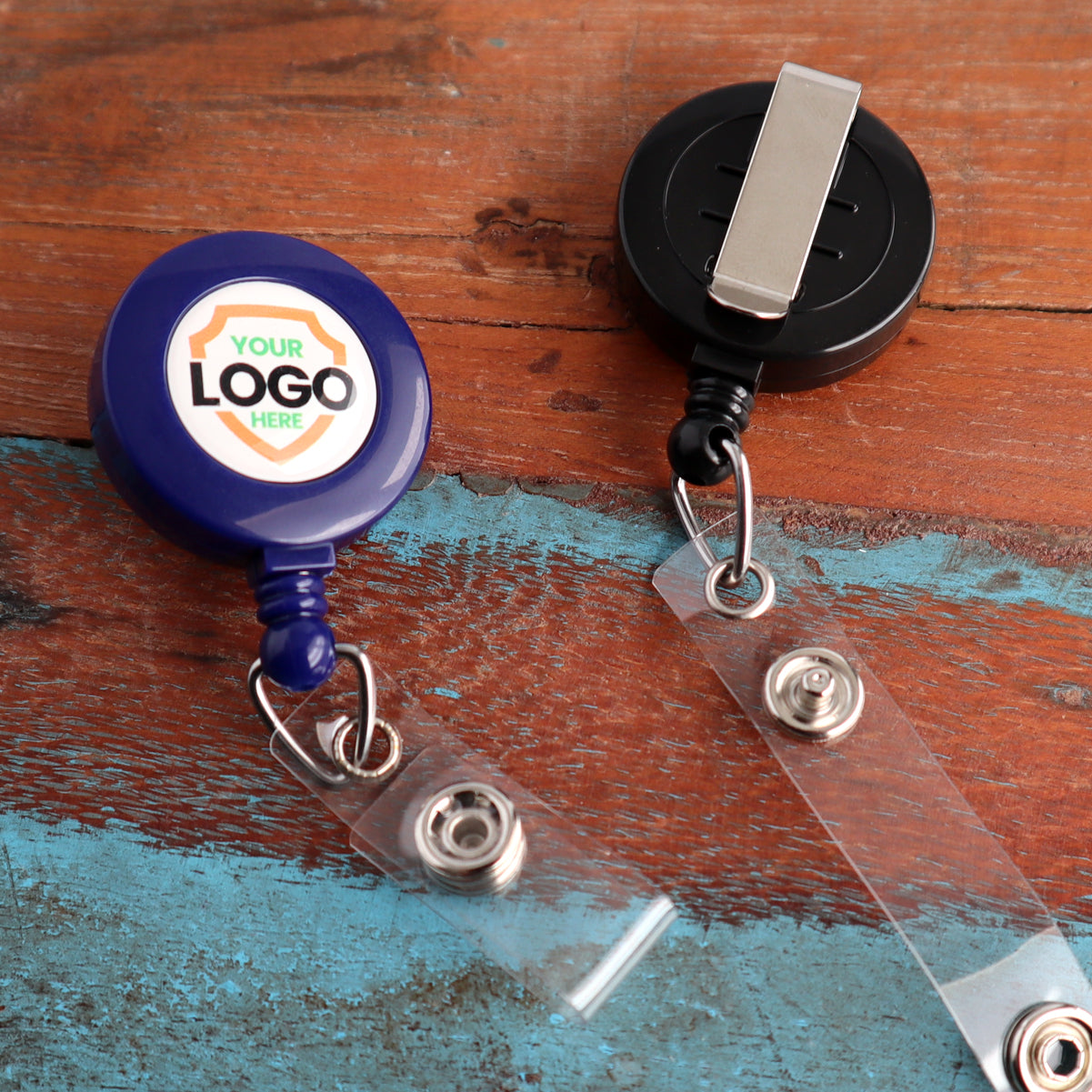 Two retractable badge holders are on a wood surface. The one on the left is Custom Printed Retractable Badge Reels With Belt Clip - Personalize with Your Brand Logo, which is blue with a customizable logo area, perfect for promoting brand awareness with full color graphics. The one on the right is black with a metal clip and clear strap, ideal for custom badge reels.