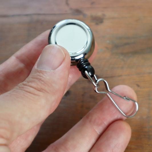 A hand holding a Mini Chrome Silver Key Reel with a spring clip and metal swivel hook against a wooden background. Sold by Specialist ID.