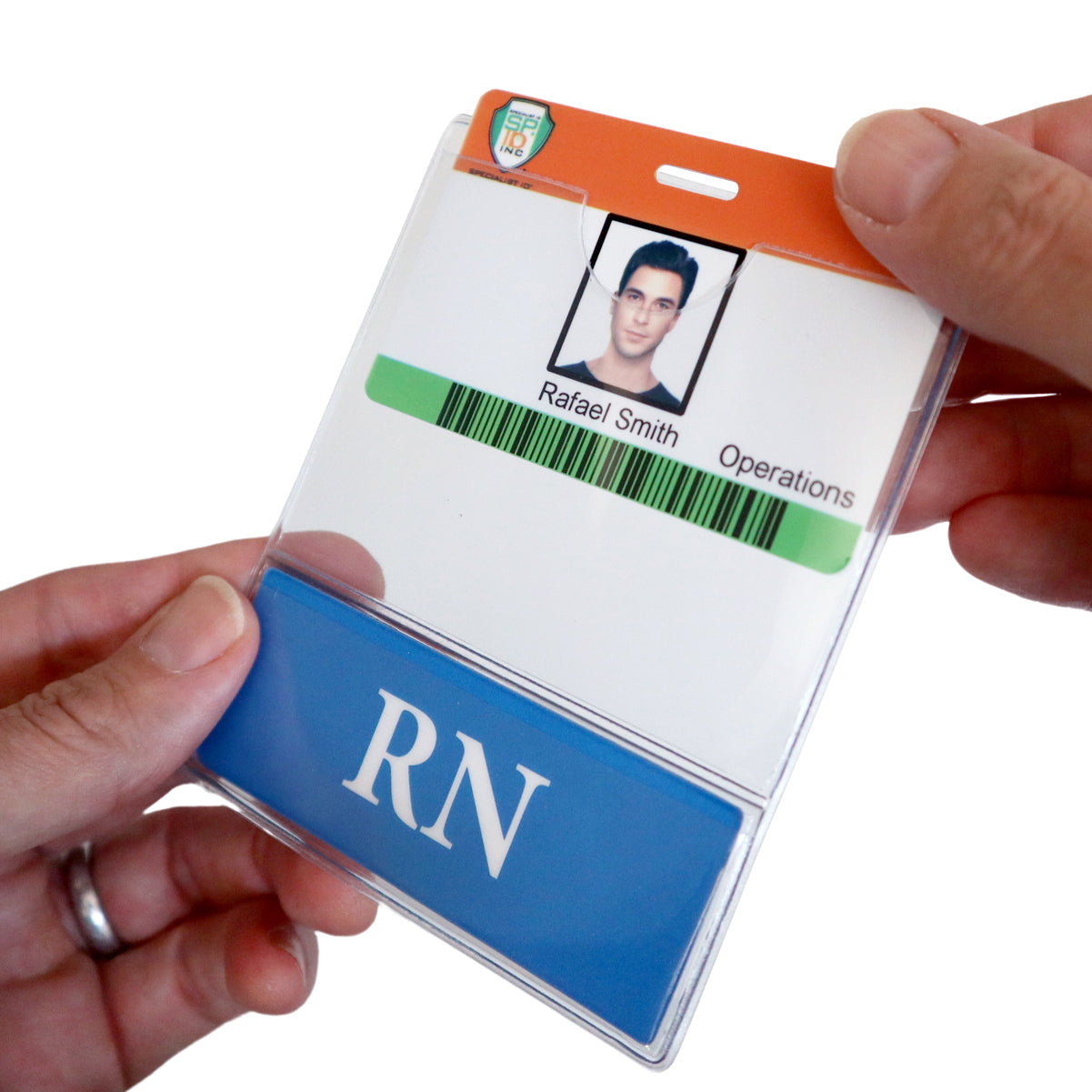 Hands holding an ID badge with a photo, the name "Rafael Smith," and the title "Operations." A blue card with "RN" is inserted in one of our RN BadgeBottom Badge Holder & RN Badge Buddy IN ONE!! - Horizontal ID Badge Sleeve with Bottom Role Tag for Registered Nurses, emphasizing professional medical titles.