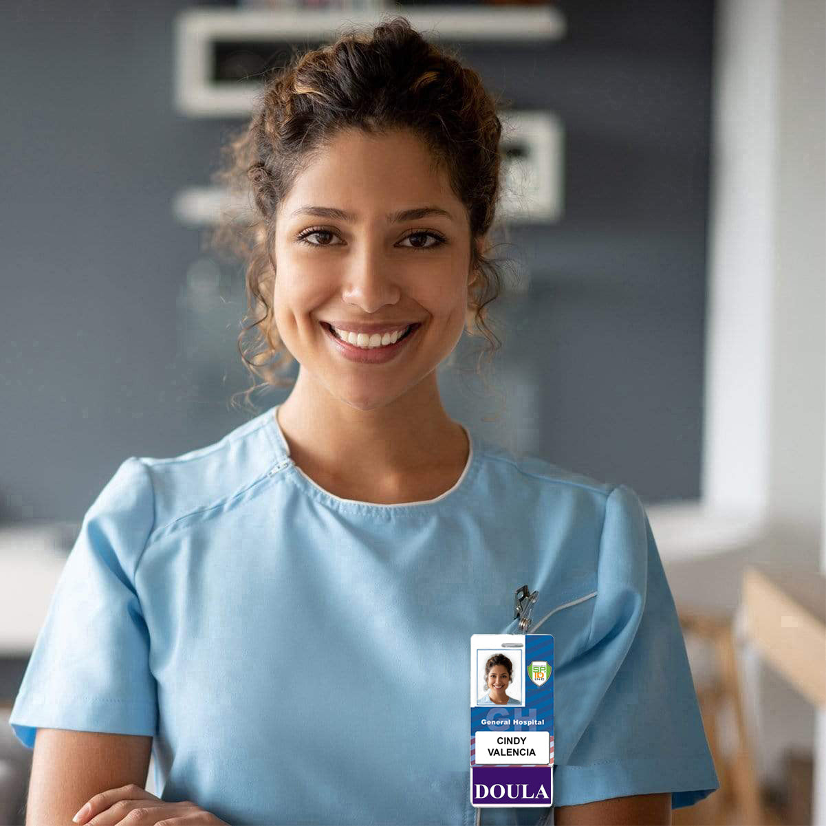 A woman wearing a blue uniform with a purple border and a DOULA Vertical Badge Buddy with Purple Border that reads "Cindy Valencia, Doula" smiles at the camera in a professional setting.