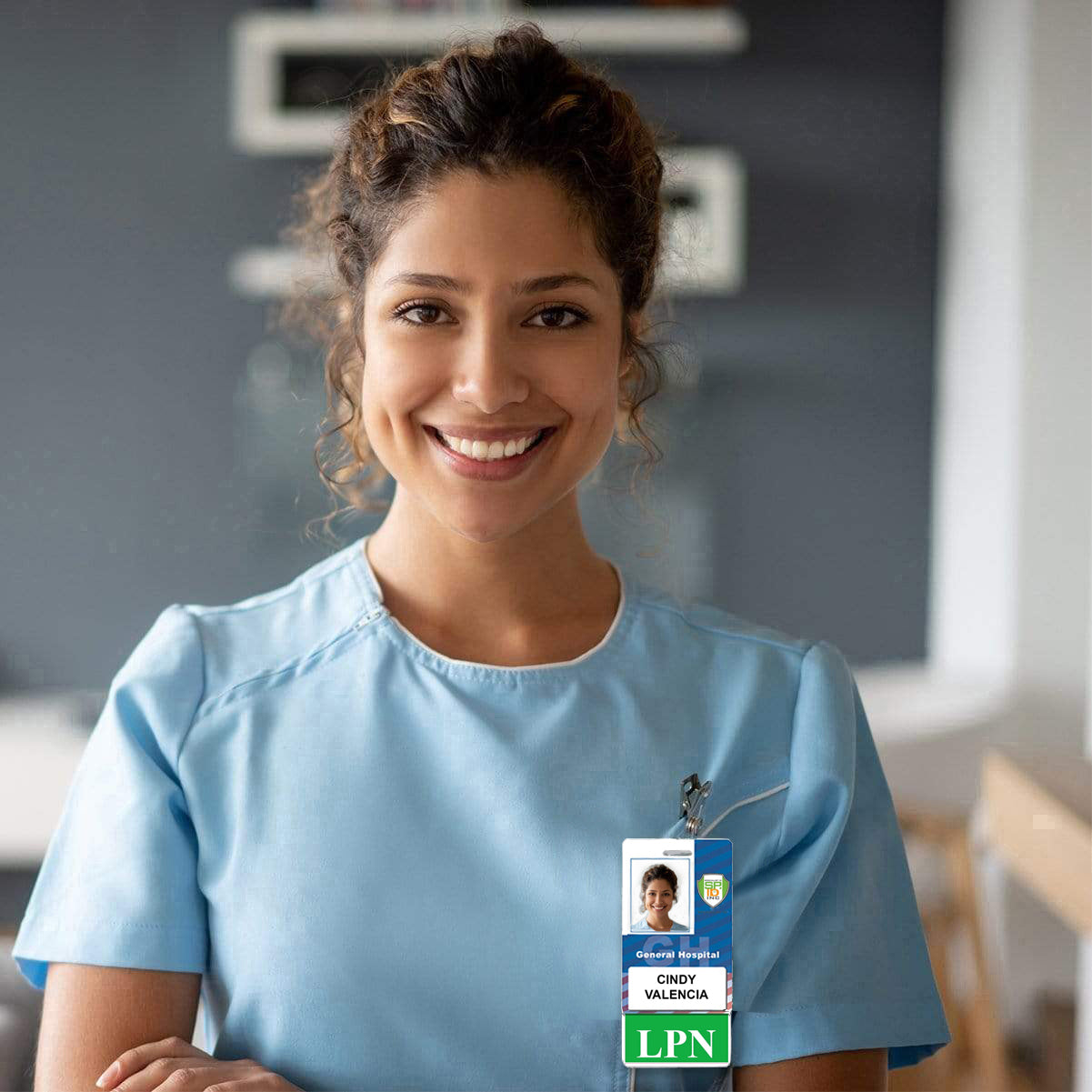 A woman wearing a light blue scrub top stands in front of a blurred background, smiling. She has an "LPN" Vertical Badge Buddy with Green Border that reads "Cindy Valencia, Licensed Practical Nurse" pinned to her uniform.