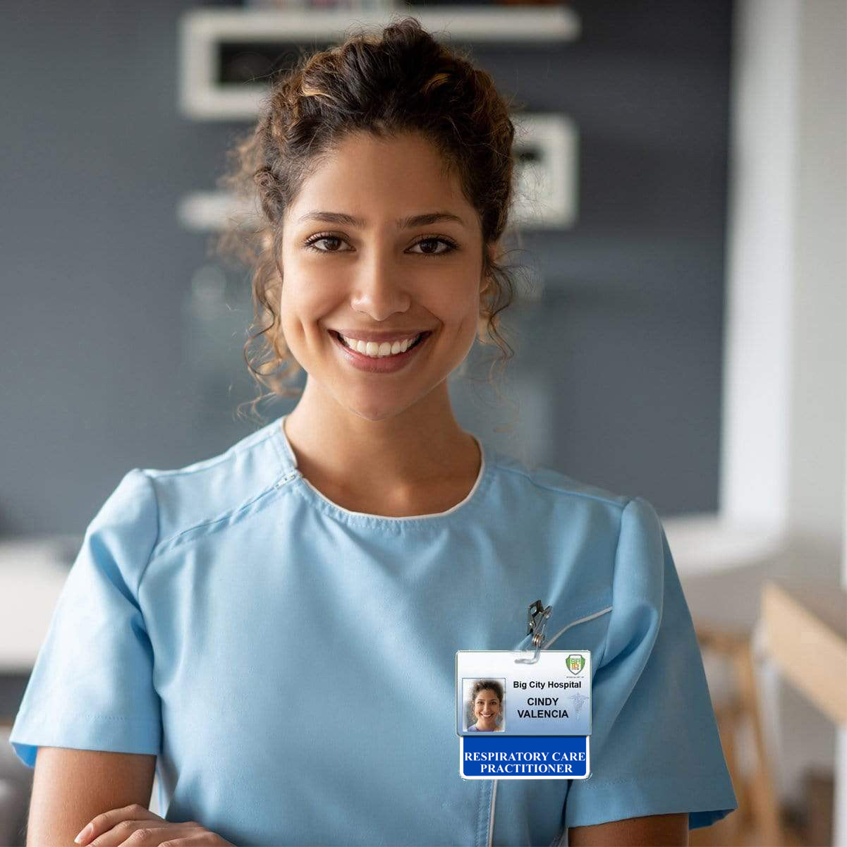 A woman in light blue scrubs, wearing a RESPIRATORY CARE PRACTITIONER Horizontal Badge Buddy with Blue Border that says "Respiratory Care Practitioner - Cindy Valencia," smiles while standing in a hospital setting.