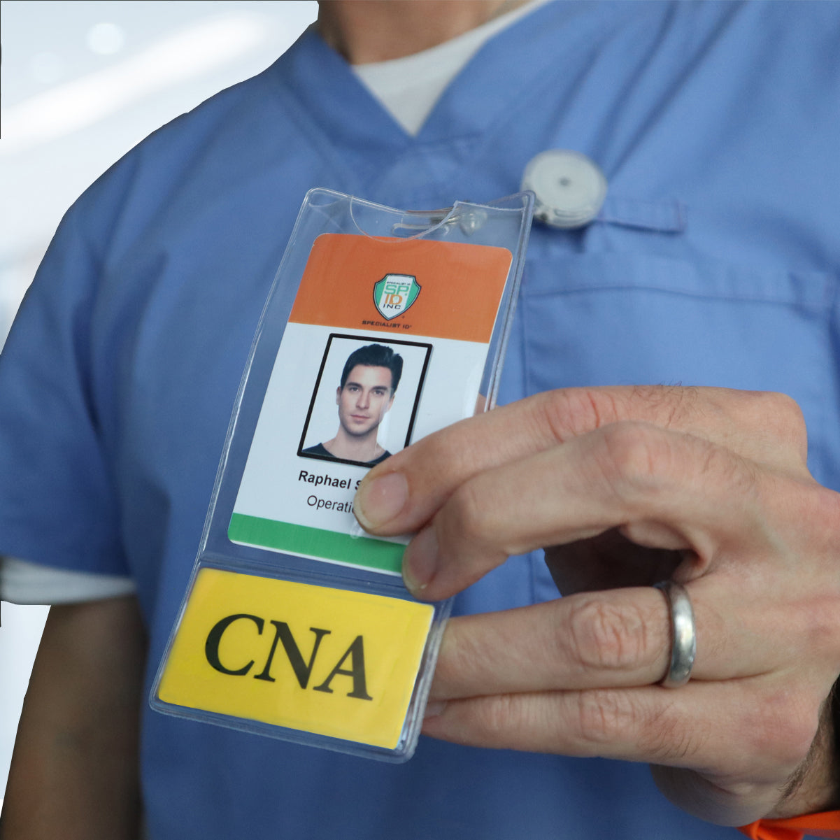 A person in blue scrubs holds an ID badge with a name and photo above a yellow card labeled "CNA," using a CNA BadgeBottom Badge Holder & Badge Buddy IN ONE!! - Vertical ID Badge Sleeve with Bottom Role Tag for Nurse Assistants.