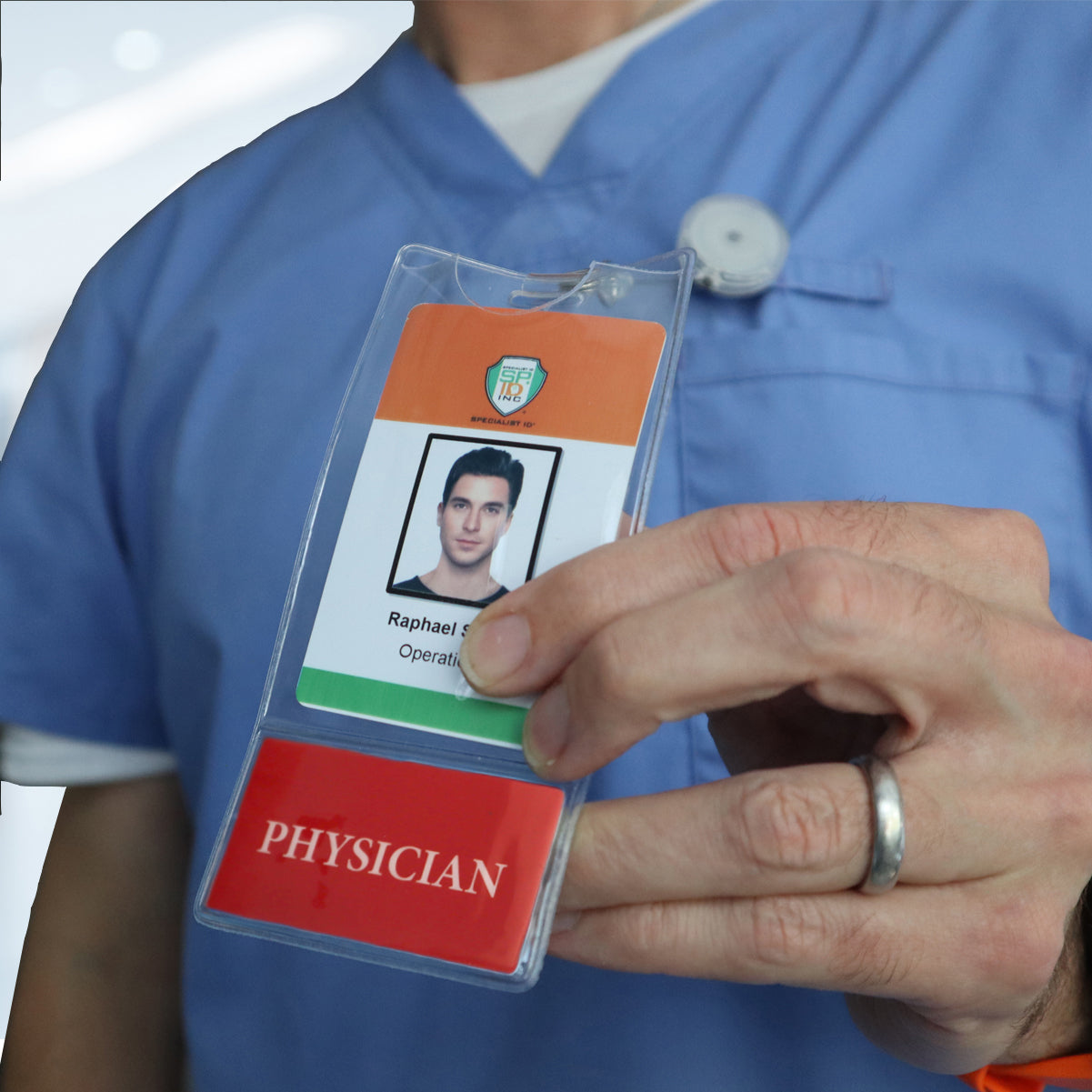 A person in a blue medical uniform holds an ID badge with the label "PHYSICIAN" and a photo of Raphael, securely displayed using the PHYSICIAN BadgeBottom Badge Holder & Badge Buddy IN ONE!! - Vertical ID Badge Sleeve with Bottom Role Tag for Doctors.