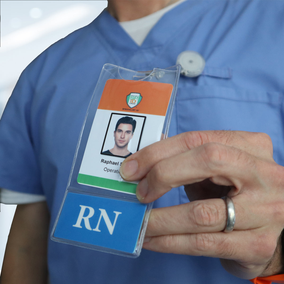A person wearing a blue uniform holds up a Custom Printed BadgeBottoms® Vertical (Badge Holder & Badge Buddy IN ONE!!) showcasing their hospital ID with a photo, name "Raphael," and title "Operations Room Nurse," with "RN" prominently displayed at the bottom.