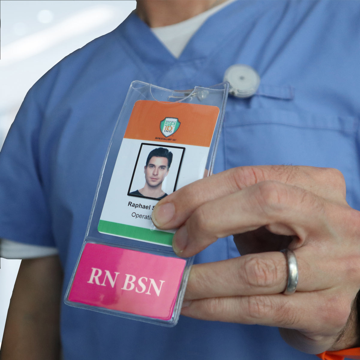 A person in blue scrubs holds an ID badge with a photo, name, and title "Operations," securely attached in one of the sleek badge holders. Beside it is a pink card labeled "RN BSN BadgeBottom Badge Holder & Badge Buddy IN ONE!! - Vertical ID Badge Sleeve with Bottom Role Tag for Registered Nurses.