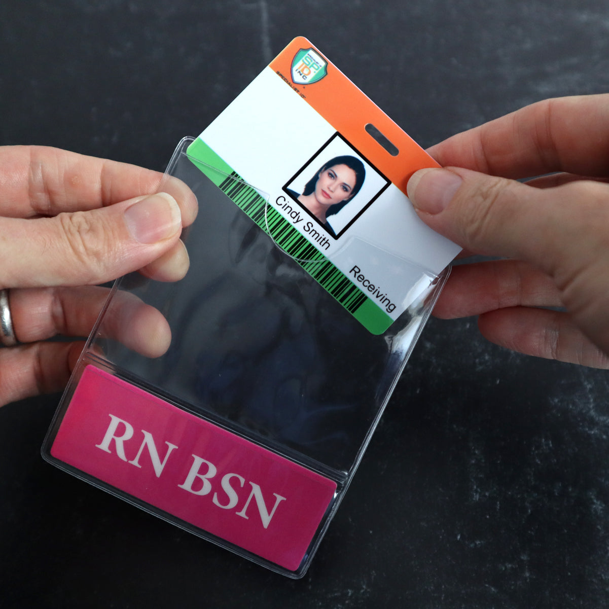 Hands holding an ID badge with a photo, the name "Cindy Smith," and the text "Receiving." Below, a label with "RN BSN" is visible. The customizable title feature and durable Custom Printed BadgeBottoms® Horizontal (Badge Holder & Badge Buddy IN ONE) complete this professional look.
