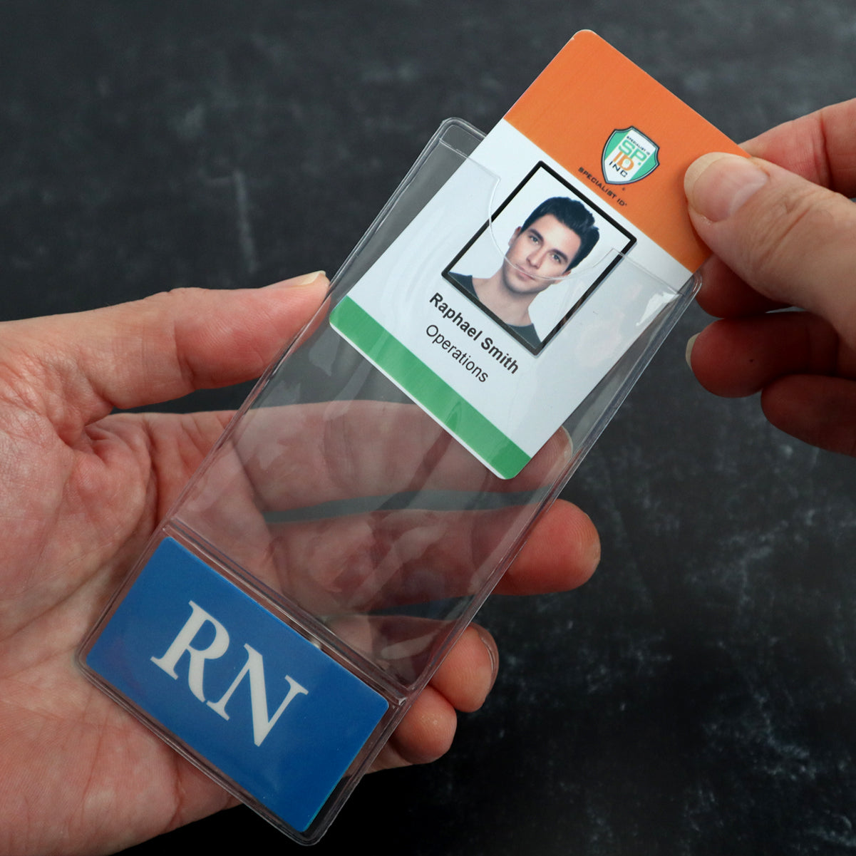 A person is inserting a badge with a photo and the text "Raphael Smith, Operations" into a Custom Printed BadgeBottoms® Vertical (Badge Holder & Badge Buddy IN ONE!!) labeled "RN.