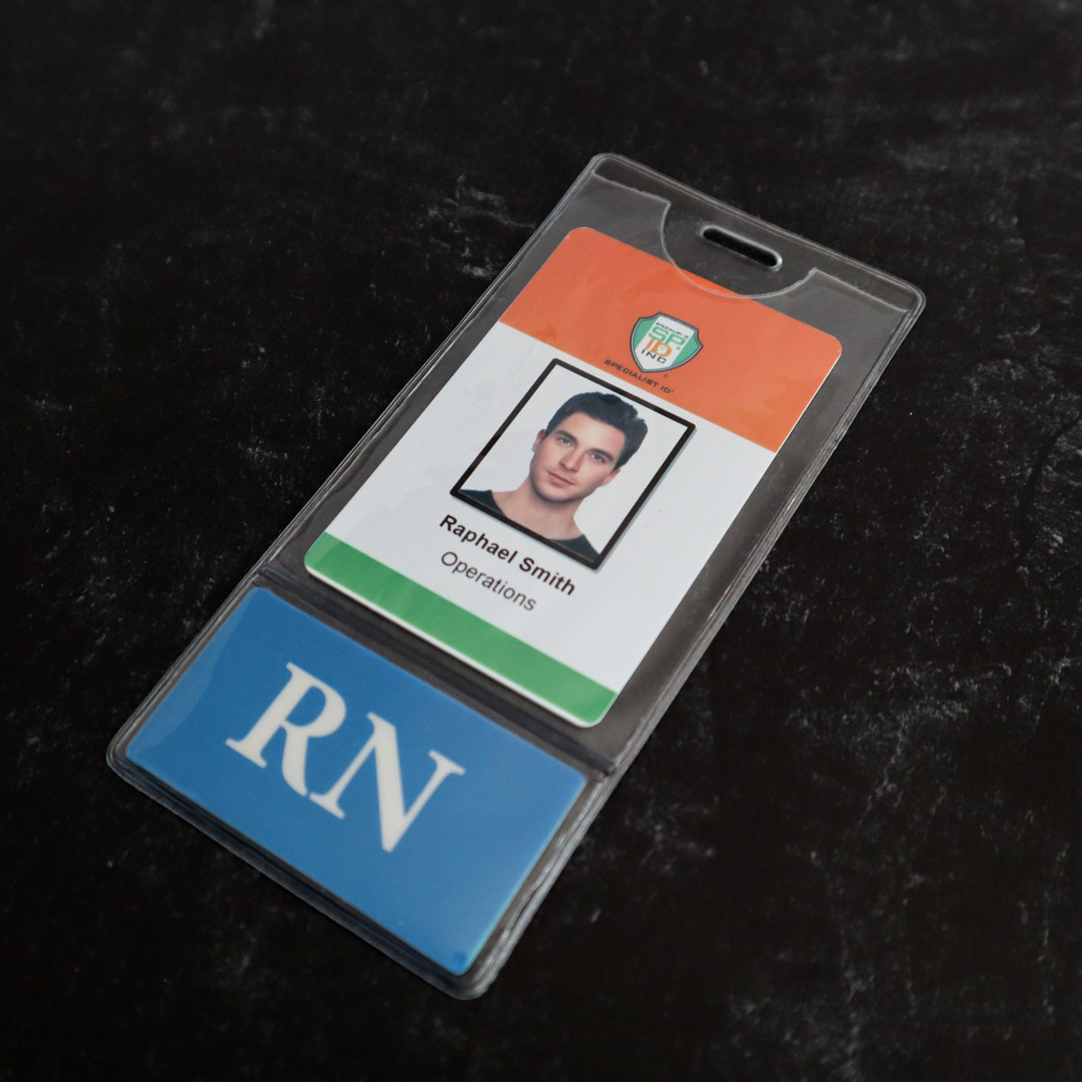 A plastic ID badge labeled “Raphael Smith, Operations” is attached to a blue card with "RN" in white letters, both encased in a clear Custom Printed BadgeBottoms® Vertical (Badge Holder & Badge Buddy IN ONE!!).