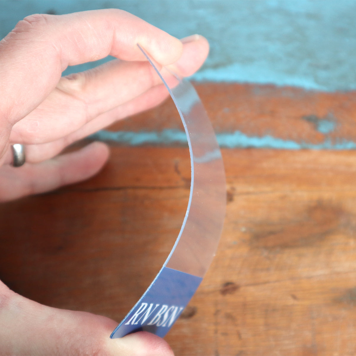 A hand bends a thin, clear, flexible Clear NURSE PRACTITIONER Badge Buddy Vertical with Blue Border for Nurses over a wooden surface. Sold by Specialist ID.