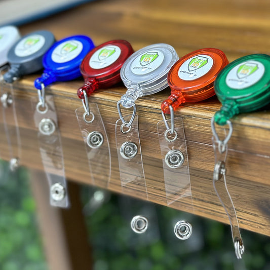 Several Translucent Custom Retractable Badge Reels With Spring Clip (2120-762X) - Add Your Logo are attached to the edge of a wooden table, perfect for enhancing brand awareness among your promotional staff.