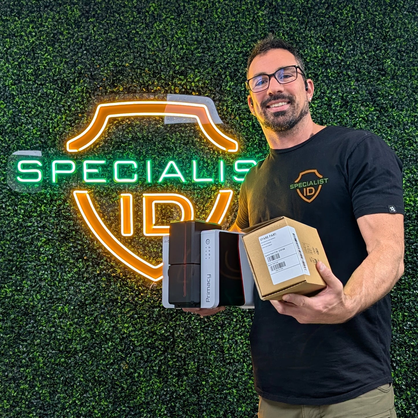 A man in glasses and a "Specialist ID" shirt stands by a neon logo, holding the Evolis S10108 Magnetic Stripe Encoder Installation Kit for Primacy & Zenius ID card printers.