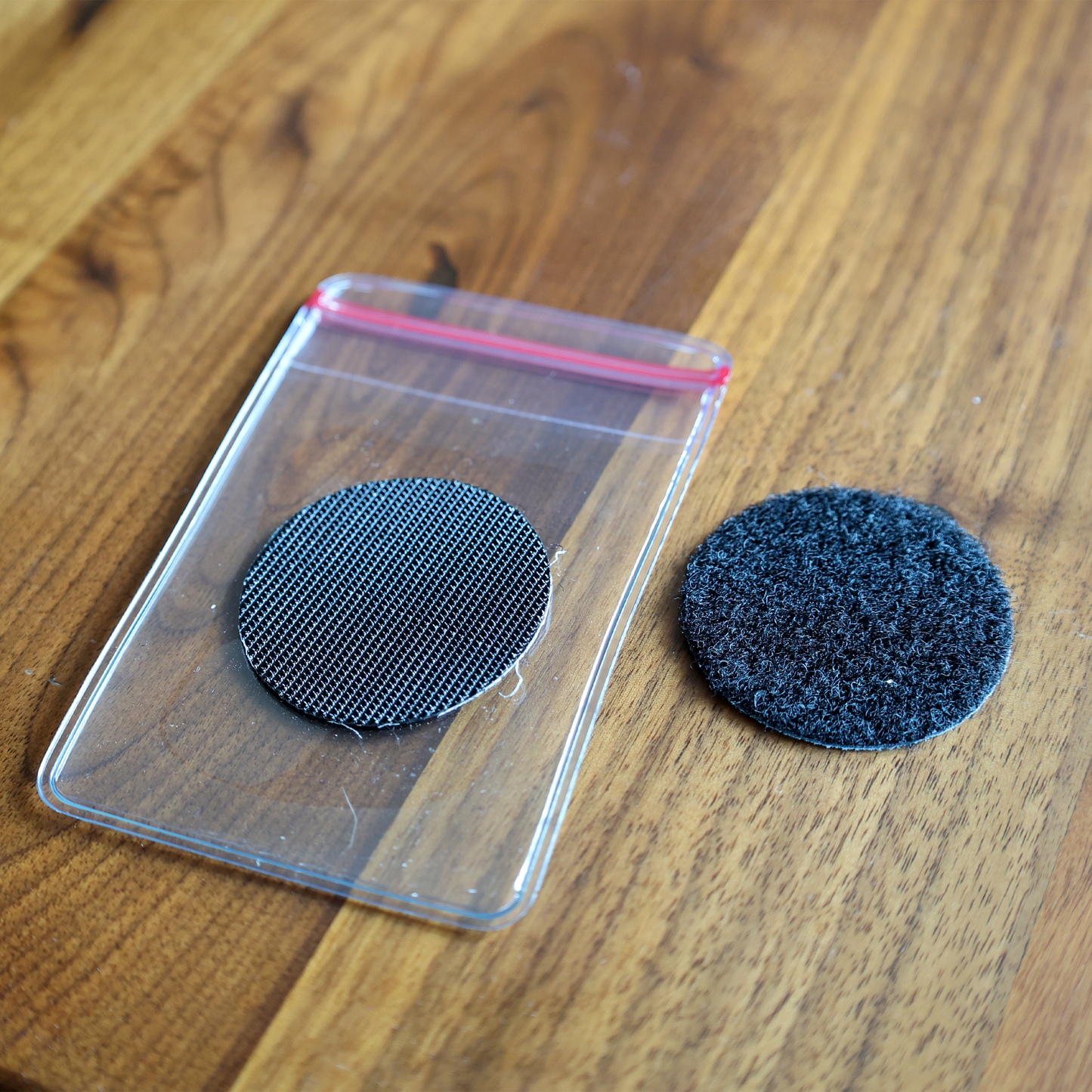 A round, textured patch is partially inside a Heavy Duty Resealable Badge Holder with Adhesive Hook & Loop Back (PPP-1), featuring a red zip. Another similar adhesive-backed patch lies on the wooden surface.