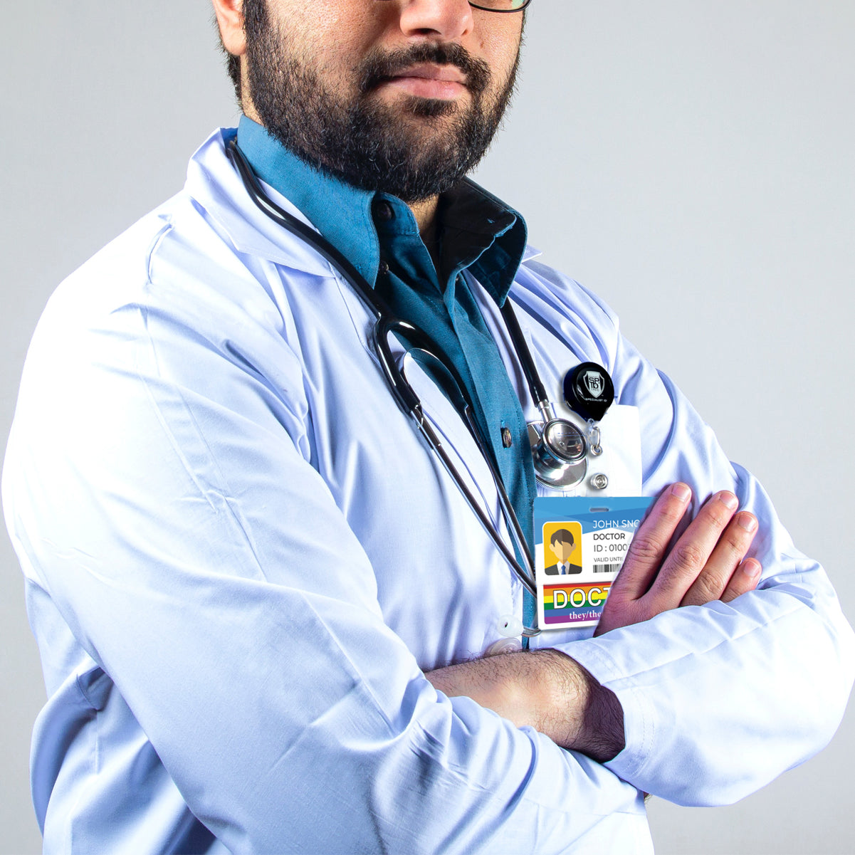 A person in a white lab coat with a stethoscope around their neck stands with arms crossed. They are wearing a blue shirt and glasses, and their ID badge reads "Doctor" with an image and text. A DOCTOR Pronouns Badge Buddy Horizontal with Rainbow Border hangs beneath their ID, adding a touch of color to their professional attire.