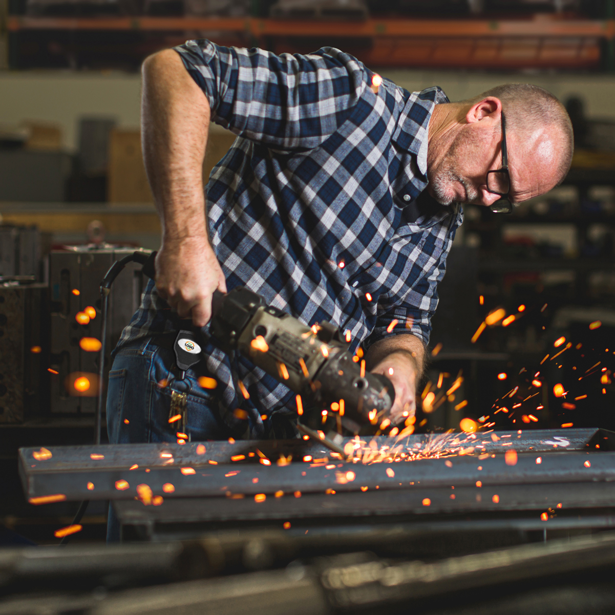 A man in a checked shirt and glasses uses a Customizable Key-Bak Super 48 Heavy Duty Key Reel (S48K) - Customize with Your Own Logo and Design to cut metal, producing sparks in a workshop setting, demonstrating rugged durability.