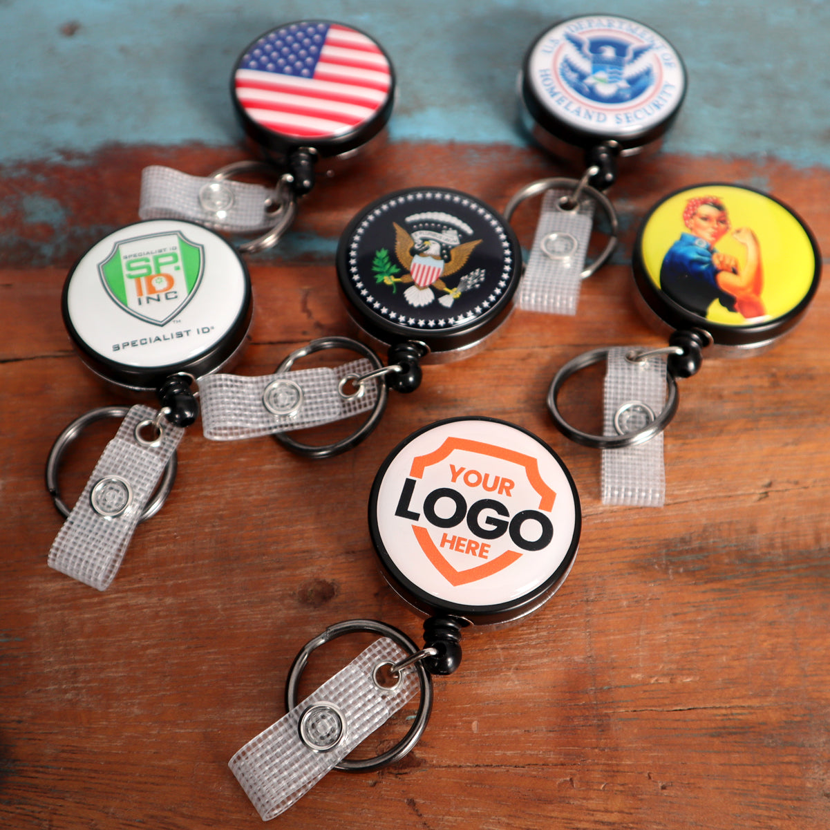 A set of six Custom Heavy Duty Badge Reel with Key Ring and Badge Strap (SPID-3180) - Add Your Logo with various designs, including a US flag, insignias, "Your Logo Here," and a "We Can Do It!" poster, featuring key rings and badge straps on a wooden surface.
