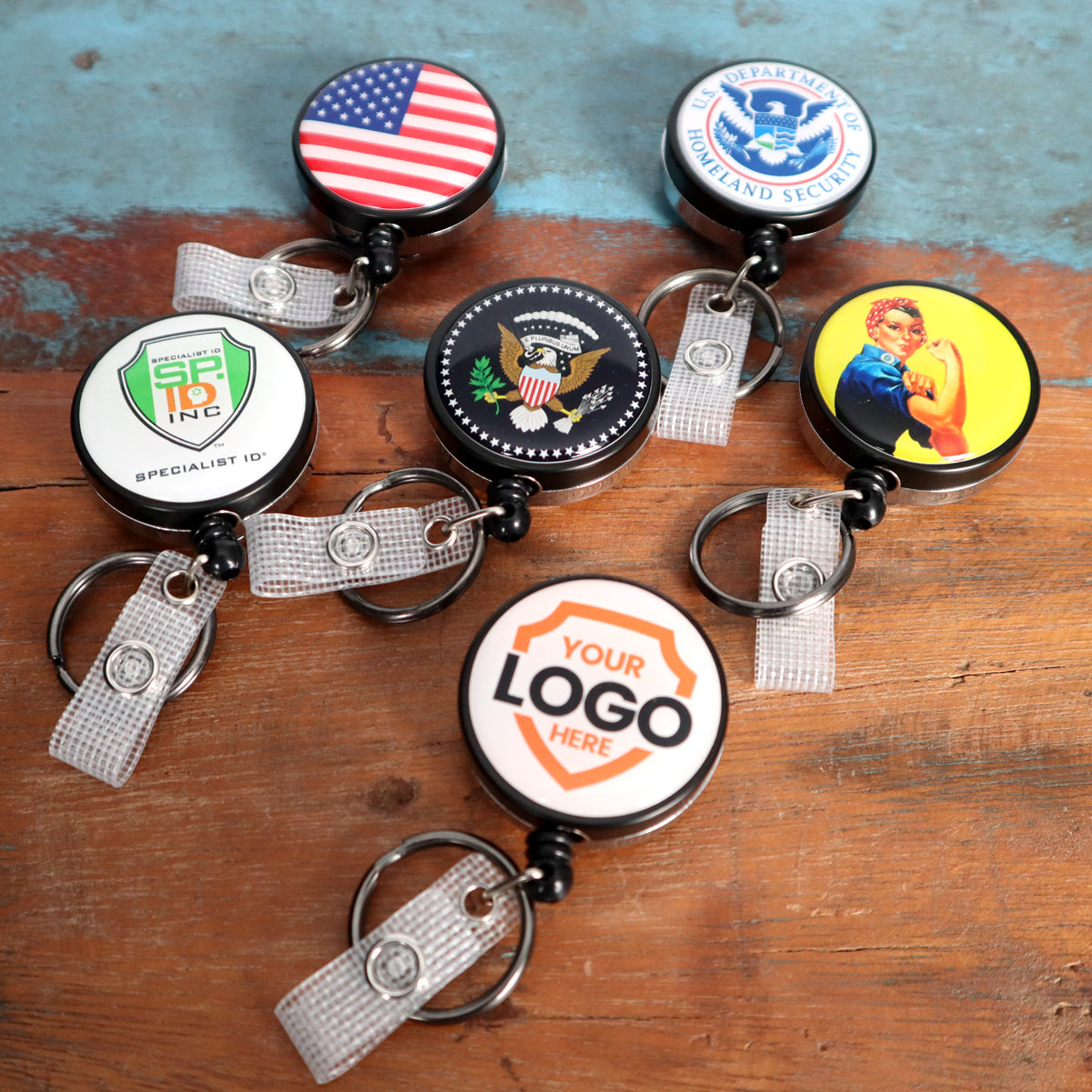 Five Custom Heavy Duty Badge Reels with Key Ring and Badge Strap (SPID-3180) - Add Your Logo with different designs, including the American flag, the Department of Homeland Security emblem, a badge with the U.S. presidential seal, "Your Logo Here," and Rosie the Riveter. Each holder features a custom heavy-duty metal badge reel and key ring for personalized identification display.