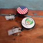 Two Custom Heavy Duty Badge Reels with Key Ring and Badge Strap (SPID-3180) - Add Your Logo rest on a wooden surface. One features a US flag design, while the other showcases the "SPECIALIST ID INC" logo. Each reel includes a key ring and badge strap for enhanced functionality and provides a personalized identification display in style.