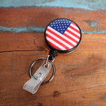 A Custom Heavy Duty Badge Reel with Key Ring and Badge Strap (SPID-3180) - Add Your Logo with an American flag design on a circular badge, attached to a small loop and key ring, placed on a wooden surface. This personalized identification display features a custom heavy-duty metal badge reel for durability.