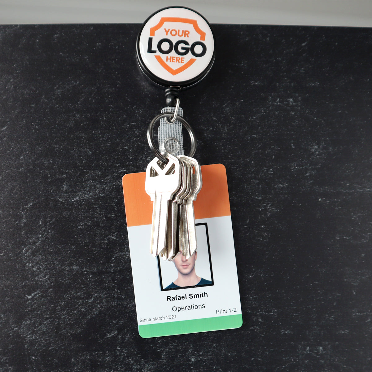 A personalized identification display featuring an employee ID badge with the name "Rafael Smith," a photo, and keys attached, is showcased against a dark background. The Custom Heavy Duty Badge Reel with Key Ring and Badge Strap (SPID-3180) - Add Your Logo adds an extra touch of durability and style.