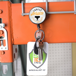 A keyring with keys, a white badge with a "Specialist ID" logo, and a round tag that reads "Carpet Service Cleaning and Repair" hangs from an orange metal shelf, securely attached to a Custom Heavy Duty Badge Reel with Key Ring and Badge Strap (SPID-3180) - Add Your Logo.