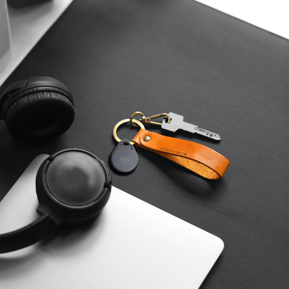 Black headphones, a laptop, and a keychain with a key and orange leather strap are placed on a black and white surface. The SPIDProx HID Compatible Key Fob enhances security through advanced access control systems.