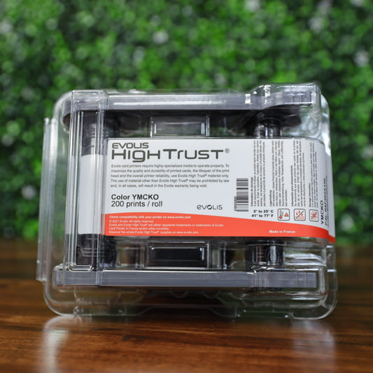 Front-facing close-up of Evolis R5F202A100 YMCKO ribbon cartridge in clear High Trust sealed packaging with visible white and red label showing 200 prints per roll for Primacy 2 ID Card Printer, placed on reflective wood surface with green foliage background. Photographed and sold by Specialist ID, an authorized Evolis dealer.