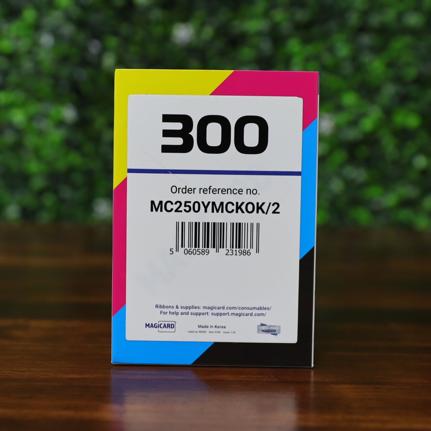 A Magicard MC250YMCKOK Ribbon–250 Dual-Sided Full-Color Prints, labeled "600" with order ref MBC250YMSCKOK/2, stands on a wooden surface against a blurred green background. The product description contains only CSS code. Photographed and sold by Specialist ID, an authorized Magicard dealer.