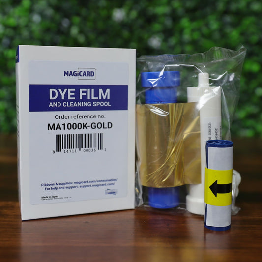 The Magicard MA1000K GOLD Gold Ribbon – 1000 Prints is displayed in packaging on a wooden surface with a blurred green background. Perfect for producing high-quality professional ID cards. Photographed and sold by Specialist ID, an authorized Magicard dealer.