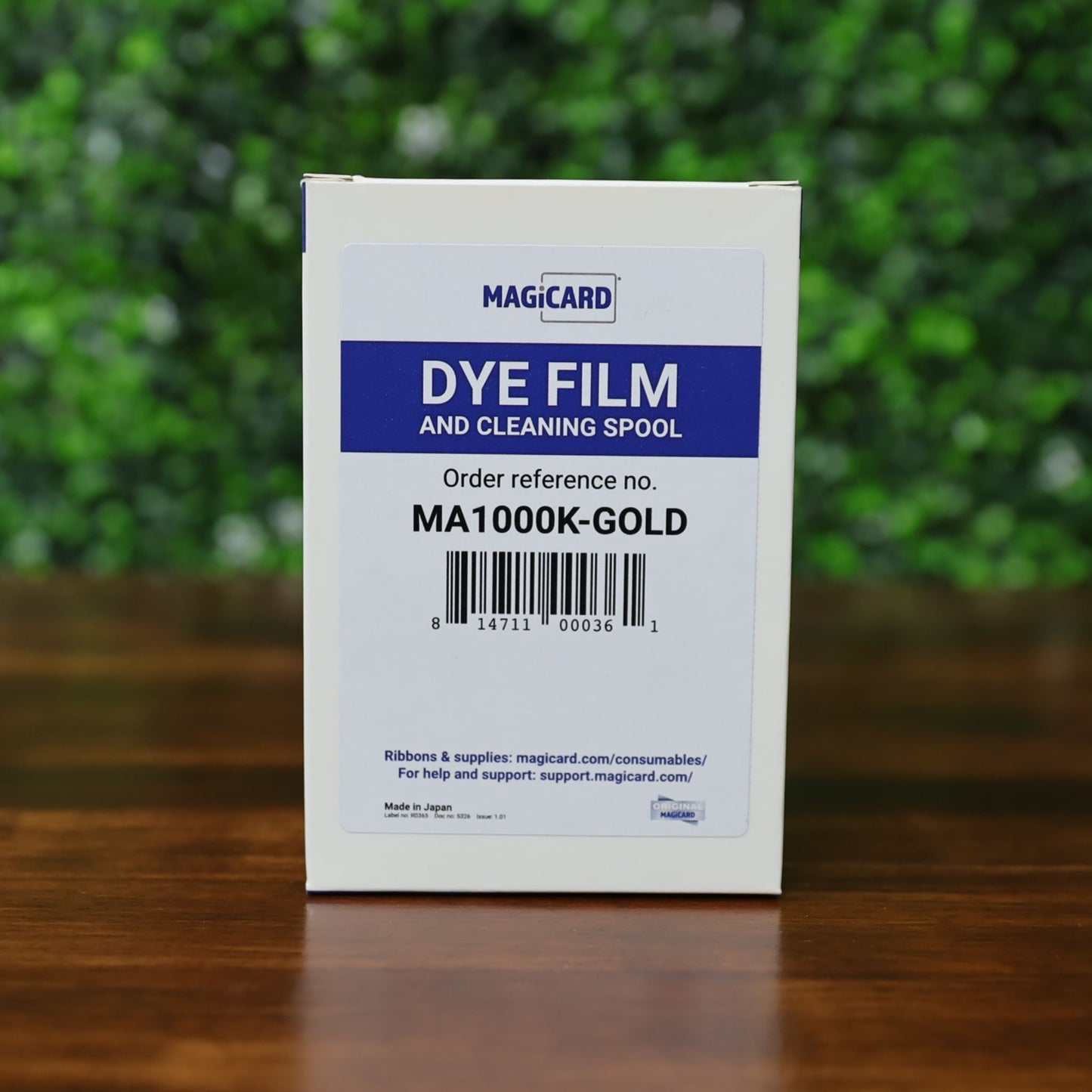 A white box labeled "Magicard MA1000K GOLD Gold Ribbon – 1000 Prints," order reference MA1000K-GOLD, is on a wooden surface with green leaves in the background—product description text is absent, replaced by CSS code. Photographed and sold by Specialist ID, an authorized Magicard dealer.