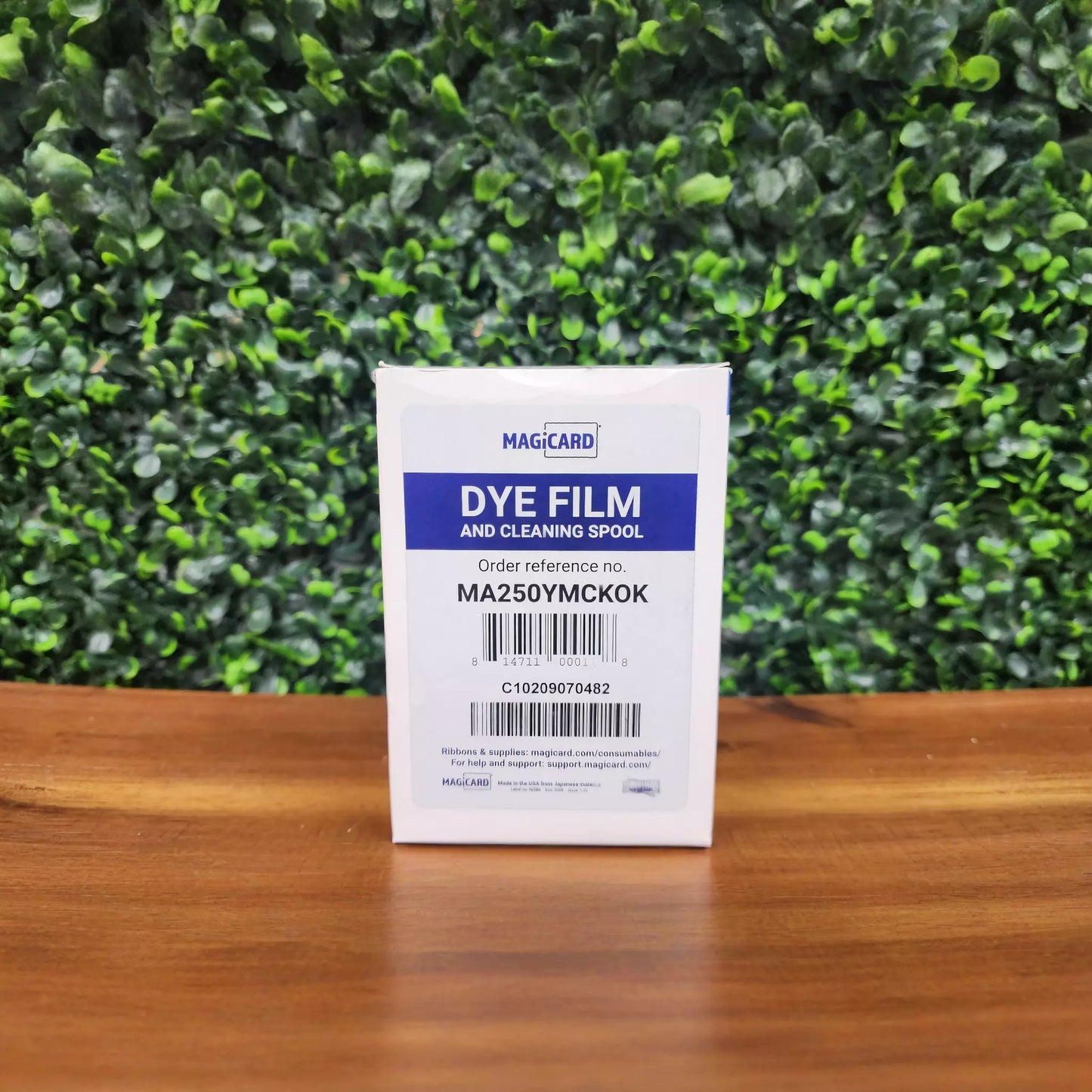 A box labeled "MAGICARD MA250YMCKOK YMCKOK Color Ribbon - 250 images" for ID card printers stands on a wooden surface with a green leafy backdrop.