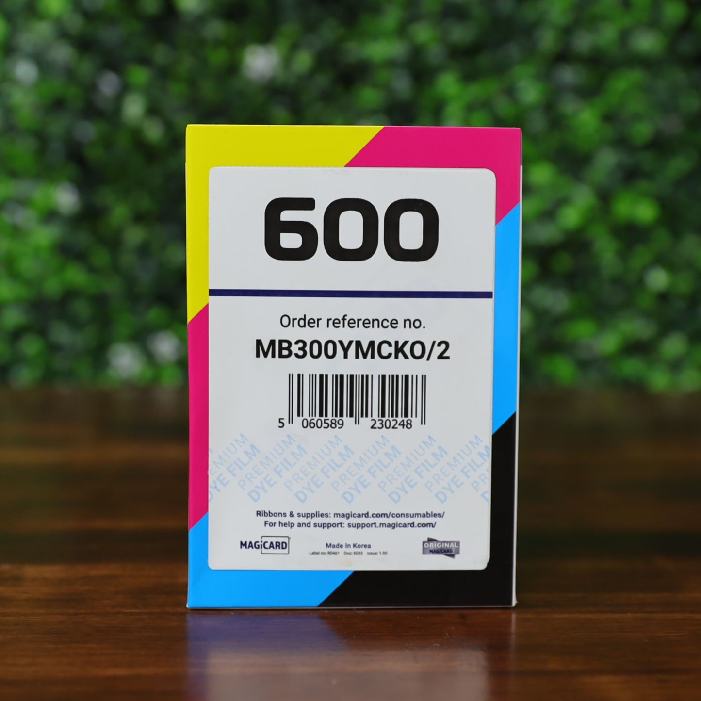 Front of a Magicard MB300YMCKOK YMCKO Ribbon box with the large “600” label, order reference MB300YMCKOK/2, barcode, and cyan, magenta, yellow, and black color stripes along the edges, placed on a wooden surface with a blurred green leafy background. Photographed and sold by Specialist ID, an authorized Magicard dealer.