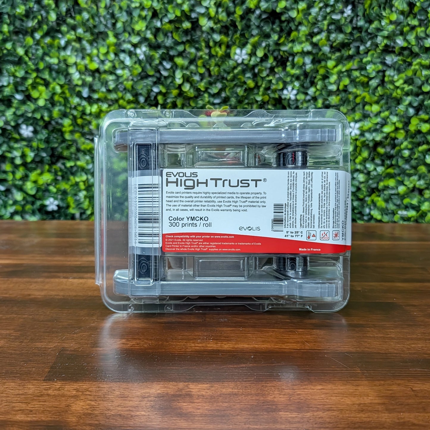 An Evolis R5F208A100 YMCKO Color Ribbon—Vibrant Full-Color Prints with Overlay Protection for the Primacy 2 ID Card Printer (300 prints/roll)—rests on a wooden surface with green leafy background, perfect for photo-quality ID card printing.