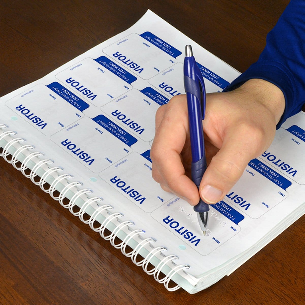 A person is meticulously writing on a sheet of Expiring Visitor Badge and Log Book - 480 Badges (05741) using a blue pen, ensuring accurate entries for the visitor management system.