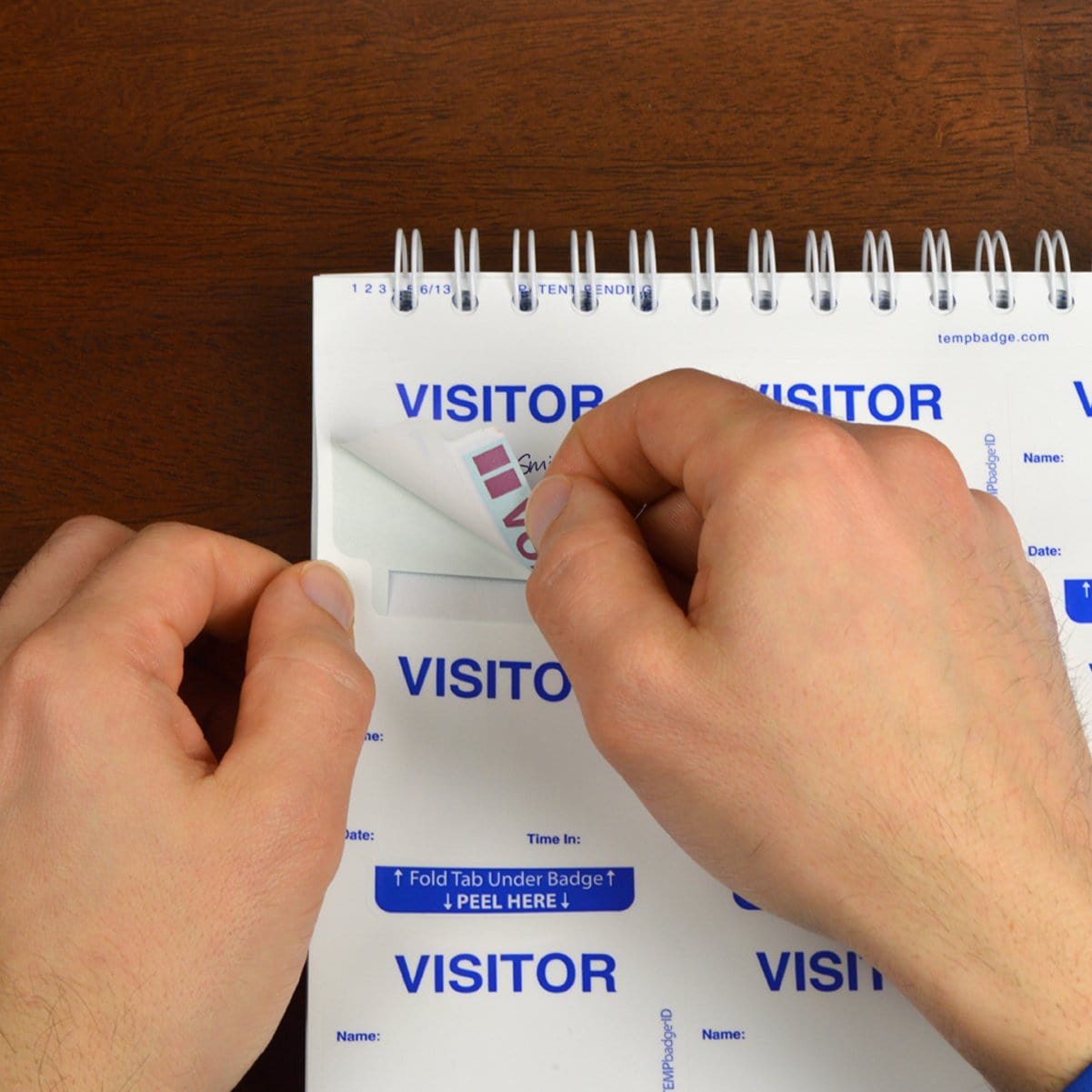 Hands peeling an Expiring Visitor Badge and Log Book - 480 Badges (05741) off a sheet of badges within a spiral-bound log book.
