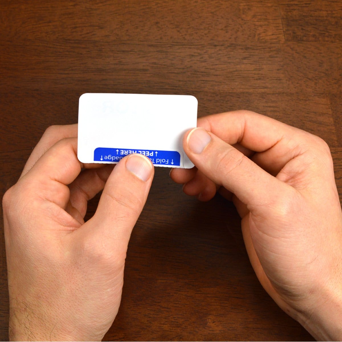 Hands holding a small rectangular white and blue card with instructions to fold and peel, designed as the Expiring Visitor Badge and Log Book - 480 Badges (05741).