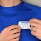 A person wearing a blue shirt is attaching an Expiring Visitor Badge and Log Book - 480 Badges (05741) to their chest that reads "Name: Edward Smith, March 25th, Time in: 9:35 AM," efficiently noted by the visitor management system.