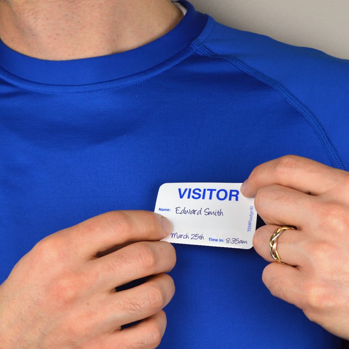A person wearing a blue shirt is attaching an Expiring Visitor Badge and Log Book - 480 Badges (05741) to their chest that reads "Name: Edward Smith, March 25th, Time in: 9:35 AM," efficiently noted by the visitor management system.