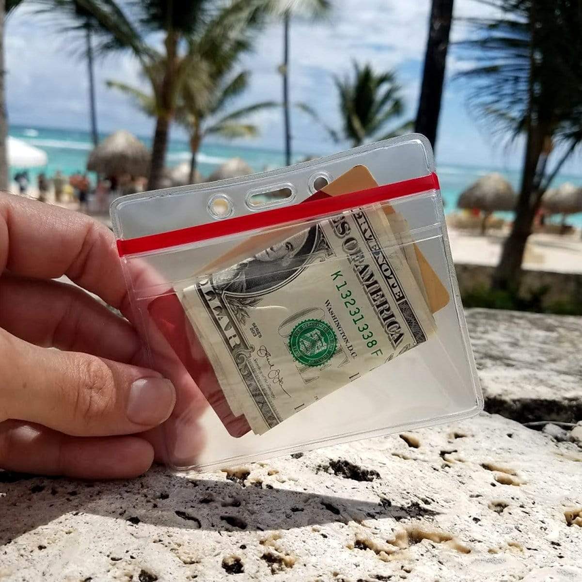 A hand holds a Heavy Duty Clear Vinyl Horizontal Badge Holder With Resealable Zip Top (1815-1010) containing a folded one-dollar bill and two cards, with a beach and palm trees in the background.