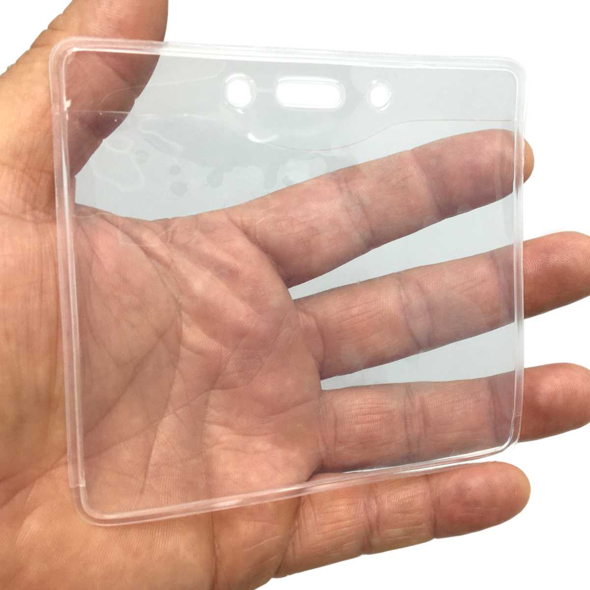 A hand holding a Clear PureClear Horizontal 4X3 Badge Holder (P/N 1815-1125), clear plastic with two holes and a slot at the top for attachment, perfect for credit card size photo IDs.