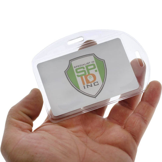 A hand holding a transparent Vertical / Horizontal Durable Clear Vinyl ID Badge Holder (1815-1600) with a green and white card inside. The card, encased in durable vinyl sleeves, displays the logo "Specialist ID, Inc." Perfect for a professional workplace environment.