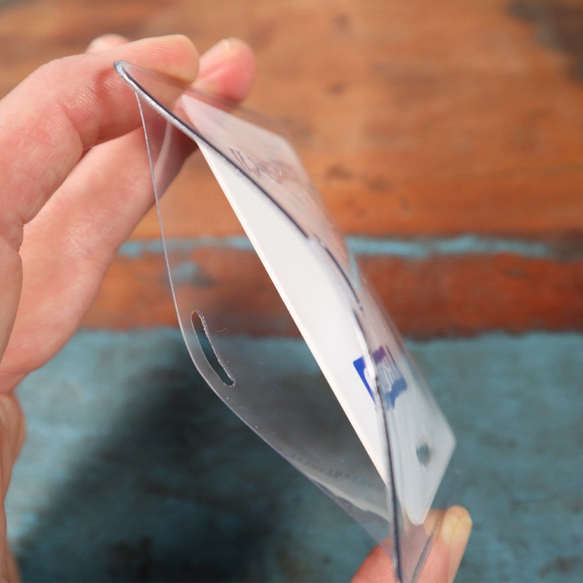 A hand holds a Clear Heavy Duty Vinyl Horizontal Proximity Card Holder 1840-5017 with a card inside, tilted to show the side view. The horizontal proximity card holder has a slot for attaching to a lanyard or clip. The background is a wooden surface.