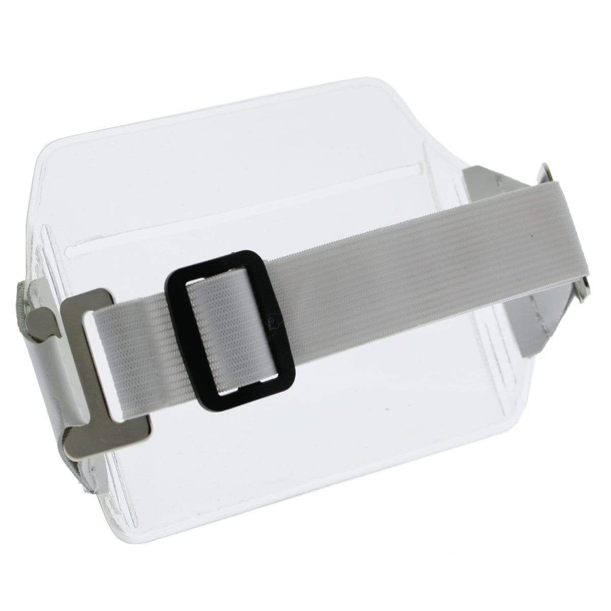 A Clear Over Size Vinyl Horizontal Arm Band Badge Holder (P/N 1840-7100) with a white adjustable strap and a black buckle is attached to a transparent rectangular plastic sheet, serving as an armband badge holder. The strap features a metal clip on one end.
